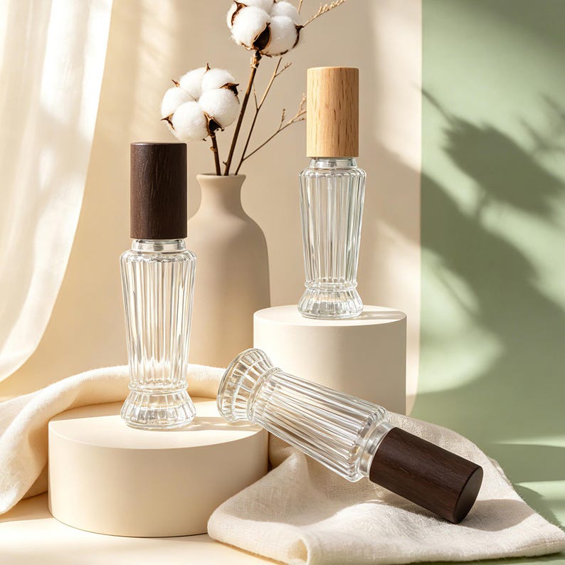 May include: Three clear glass spray bottles with vertical ribbed textures. Two bottles feature dark brown wooden caps, while one bottle has a light tan wooden cap. The bottles are arranged on cylindrical platforms with a soft fabric drape.