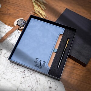 May include: A blue leather journal with the name "Alexander" and a decorative "A" initial is presented in a black gift box. A black and gold pen rests inside the box. A brown leather watch with a black face is visible in the background.
