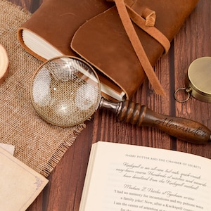 May include: A magnifying glass with a dark brown wooden handle engraved with the name Alexander, featuring a brass-toned rim and a clear glass lens. The handle has a ribbed texture and a rounded end, positioned near a leather-bound journal.