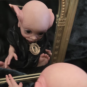 May include: A doll with pale skin, large eyes, and pointed ears, dressed in a black robe and a gold pendant. The doll is reflected in a mirror, with a hand reaching towards it. The doll's features are detailed, creating a unique and artistic appearance.