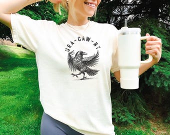 May include: Cream-colored t-shirt with a black raven graphic and the text "URA-CAW-NT" in a circle. A large white tumbler with a handle is held in the hand. The background features green foliage.