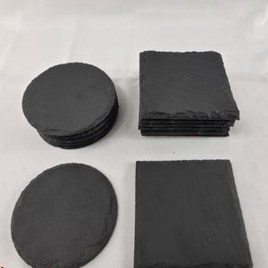 Puede incluir: Una colección de posavasos de pizarra negra con formas circulares y cuadradas. El conjunto incluye una pila de posavasos redondos, una pila de posavasos cuadrados, un posavasos redondo individual y uno cuadrado individual, todos con bordes rugosos.