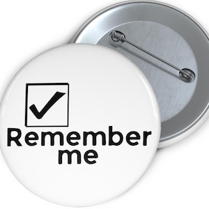 Puede incluir: Botón blanco con el texto "Remember me" en negro. Una marca de verificación negra está dentro de un cuadrado negro. El botón tiene un respaldo de alfiler plateado.
