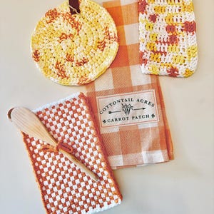 May include: A collection of kitchen textiles and accessories in shades of orange, yellow, and white. Includes a round crocheted trivet, a rectangular crocheted item, a checkered dish towel, and a woven pot holder with a wooden spoon. The dish towel has the text "COTTONTAIL ACRES CARROT PATCH".