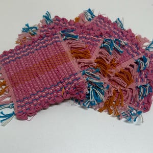 May include: A stack of four square woven coasters. The coasters are primarily pink with blue, orange, and white fringe. The weave pattern is visible, and the coasters are stacked on a white surface.
