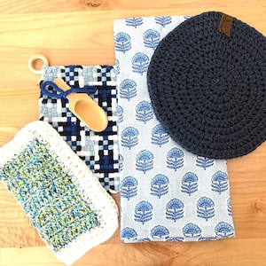 May include: A collection of kitchen textiles and accessories, including a navy blue crocheted hot pad, a blue and white patterned dish towel, a woven pot holder with a wooden scoop, and a crocheted washcloth with a white border.