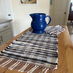 May include: A cobalt blue ceramic pitcher with a handle sits on a woven runner with fringe. The runner features horizontal stripes in shades of blue, black, and beige. The pitcher is on a wooden table.