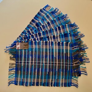 May include: A stack of handmade woven placemats in shades of blue, teal, gray, and white. Each placemat has a fringed edge. The placemats are arranged in a fan shape, showcasing the vertical stripe pattern and color variations.