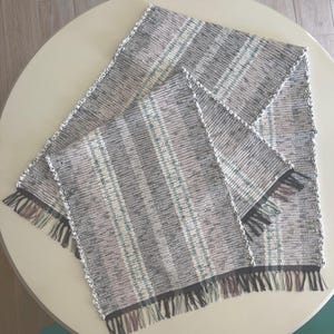 May include: A rectangular woven table runner featuring a striped pattern in shades of gray, white, and teal. The textile has a textured weave with visible vertical lines and decorative fringe detailing along the short ends of the fabric.