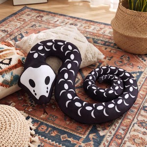 May include: A long, plush snake-shaped cushion in black with a white head and a repeating white crescent moon pattern along its body. The snake has a pointed white head with black eye markings and a small, dark brown tongue protruding from the mouth.