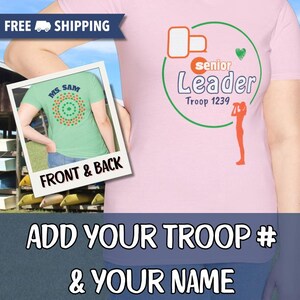 May include: A light pink t-shirt with a graphic design for a Senior Leader of Troop 1239. The design includes a green circle, orange and green accents, and the text "ADD YOUR TROOP # & YOUR NAME". A smaller image shows the back of a green shirt.