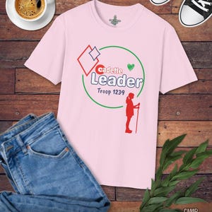 May include: A light pink t-shirt with the words "Cadette Leader Troop 1239" in a graphic design. The shirt is paired with blue jeans, black sneakers, and a cup of coffee on a wooden surface.