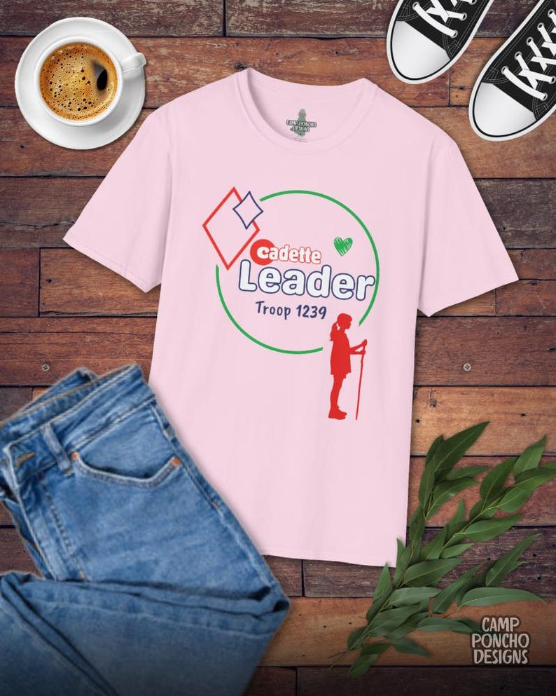 May include: A light pink t-shirt with the words "Cadette Leader Troop 1239" in a graphic design. The shirt is paired with blue jeans, black sneakers, and a cup of coffee on a wooden surface.