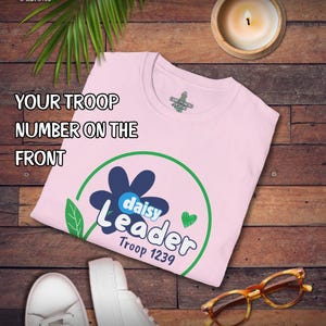 May include: A light pink t-shirt with a daisy design and the words "Daisy Leader Troop 1239" in white and blue. The shirt is on a wooden surface with a pair of white sneakers, glasses, and a lit candle.