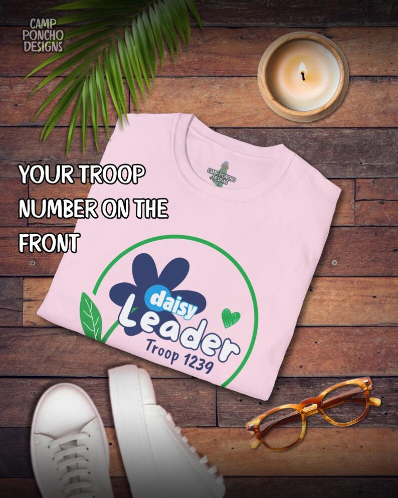 May include: A light pink t-shirt with a daisy design and the words "Daisy Leader Troop 1239" in white and blue. The shirt is on a wooden surface with a pair of white sneakers, glasses, and a lit candle.