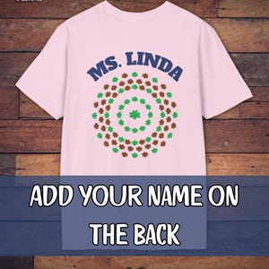 May include: A light pink t-shirt with the text "MS. LINDA" in navy blue above a circular design of green and brown shamrocks. Below, the text "ADD YOUR NAME ON THE BACK" is displayed. The Camp Poncho Designs logo is in the top left corner.