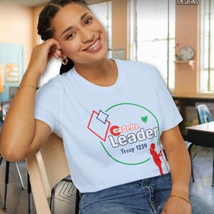 May include: White t-shirt with a graphic that says "Cadette Leader Troop 1239" in red, green, and white. The shirt features a red and blue geometric design and a silhouette of a person. The shirt is paired with blue jeans.