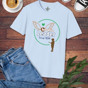 May include: A light blue t-shirt with a graphic that reads "Brownie Leader Troop 1239" in green and brown. The shirt is paired with blue jeans, black and white sneakers, and a cup of coffee on a wooden surface.