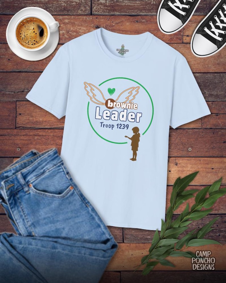 May include: A light blue t-shirt with a graphic that reads "Brownie Leader Troop 1239" in green and brown. The shirt is paired with blue jeans, black and white sneakers, and a cup of coffee on a wooden surface.