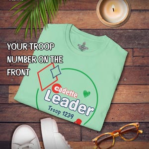May include: A light green t-shirt with the text "cadette Leader Troop 1239" in white and red. The shirt also has a red diamond, a blue diamond, and a green heart. The shirt is on a wooden surface.
