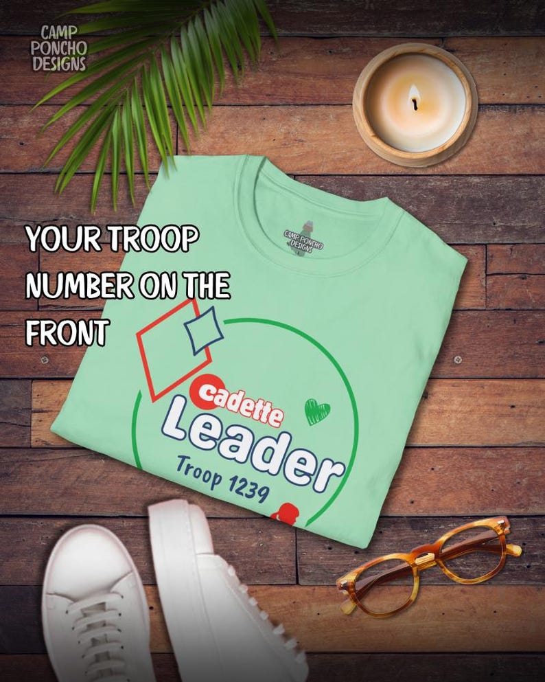 May include: A light green t-shirt with the text "cadette Leader Troop 1239" in white and red. The shirt also has a red diamond, a blue diamond, and a green heart. The shirt is on a wooden surface.