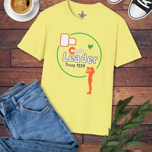 May include: A yellow t-shirt with the words "Senior Leader Troop 1239" in green and orange, with a green heart and an orange silhouette of a person. The shirt is next to blue jeans, a cup of coffee, and black and white sneakers.