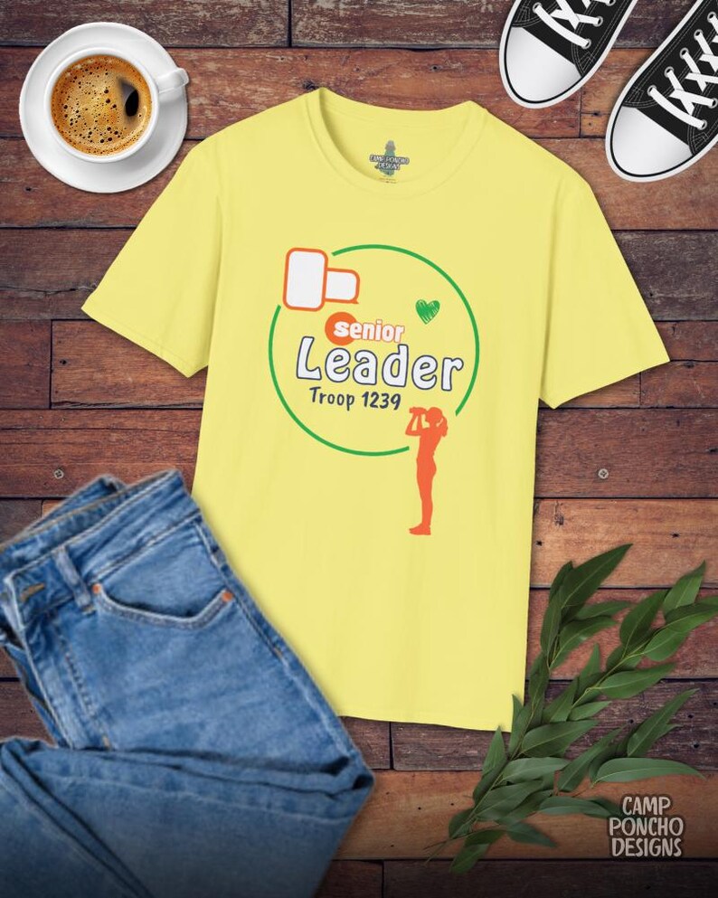 May include: A yellow t-shirt with the words "Senior Leader Troop 1239" in green and orange, with a green heart and an orange silhouette of a person. The shirt is next to blue jeans, a cup of coffee, and black and white sneakers.