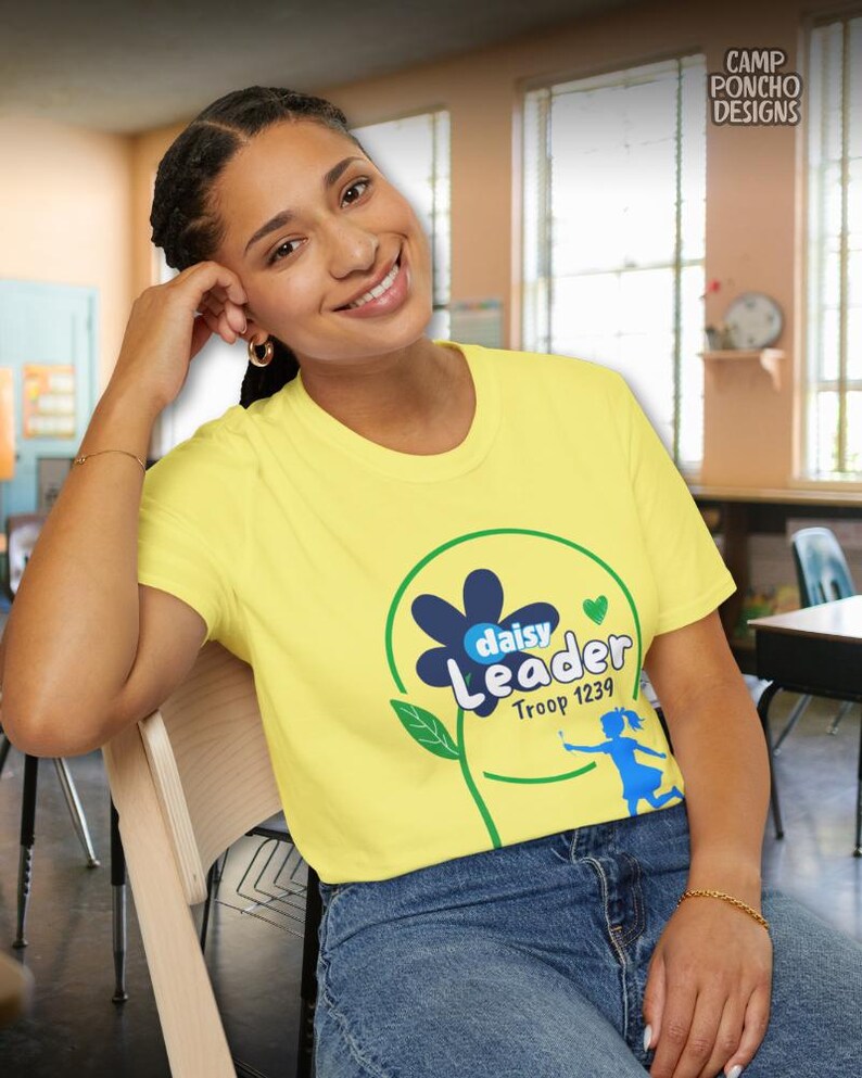 May include: A yellow t-shirt with a daisy flower design and the words "daisy Leader Troop 1239". The shirt is worn by a person in blue jeans, sitting in a classroom setting.