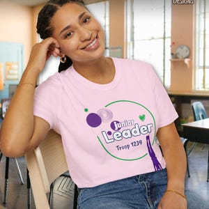 May include: A light pink t-shirt with the words "Junior Leader" in a green circle, purple and green graphics, and the text "Troop 1239". The shirt is worn with blue jeans.