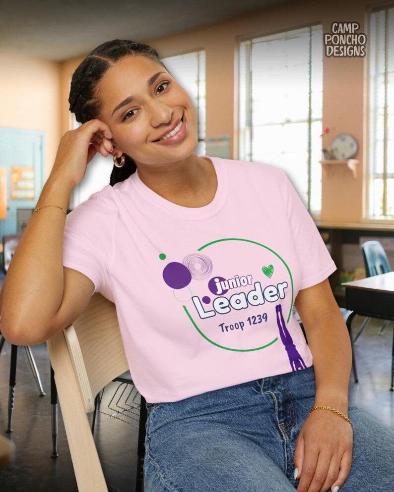 May include: A light pink t-shirt with the words "Junior Leader" in a green circle, purple and green graphics, and the text "Troop 1239". The shirt is worn with blue jeans.