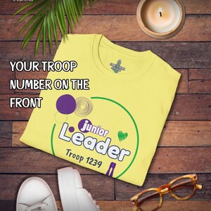 May include: A yellow t-shirt with the text "Junior Leader" and "Troop 1239" in green and purple. The shirt also has the text "Your Troop Number on the Front". The shirt is on a wooden surface with a candle, shoes, and glasses.