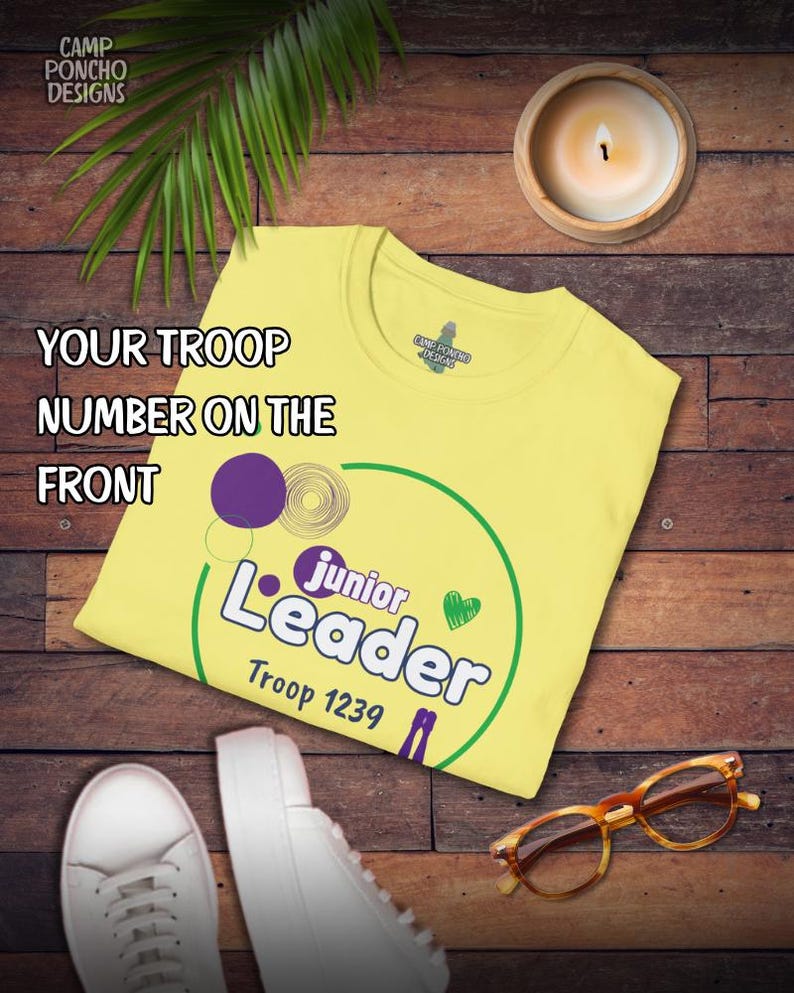 May include: A yellow t-shirt with the text "Junior Leader" and "Troop 1239" in green and purple. The shirt also has the text "Your Troop Number on the Front". The shirt is on a wooden surface with a candle, shoes, and glasses.