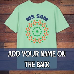 May include: A light green t-shirt with the text "MS. SAM" in blue. The shirt features a circular design of green and orange shamrocks. The text "ADD YOUR NAME ON THE BACK" is displayed below the design.