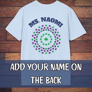 May include: Light blue t-shirt with the text "MS. NAOMI" in navy blue, above a circular design of green and purple flower shapes. The text "ADD YOUR NAME ON THE BACK" is displayed on a blue banner. A green heart is at the bottom.