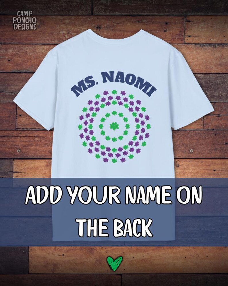 May include: Light blue t-shirt with the text "MS. NAOMI" in navy blue, above a circular design of green and purple flower shapes. The text "ADD YOUR NAME ON THE BACK" is displayed on a blue banner. A green heart is at the bottom.