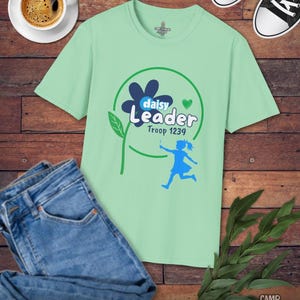 May include: A light green t-shirt with a graphic that says "Daisy Leader Troop 1239" with a flower and a running girl. The shirt is paired with blue jeans, black sneakers, and a cup of coffee on a wooden surface.