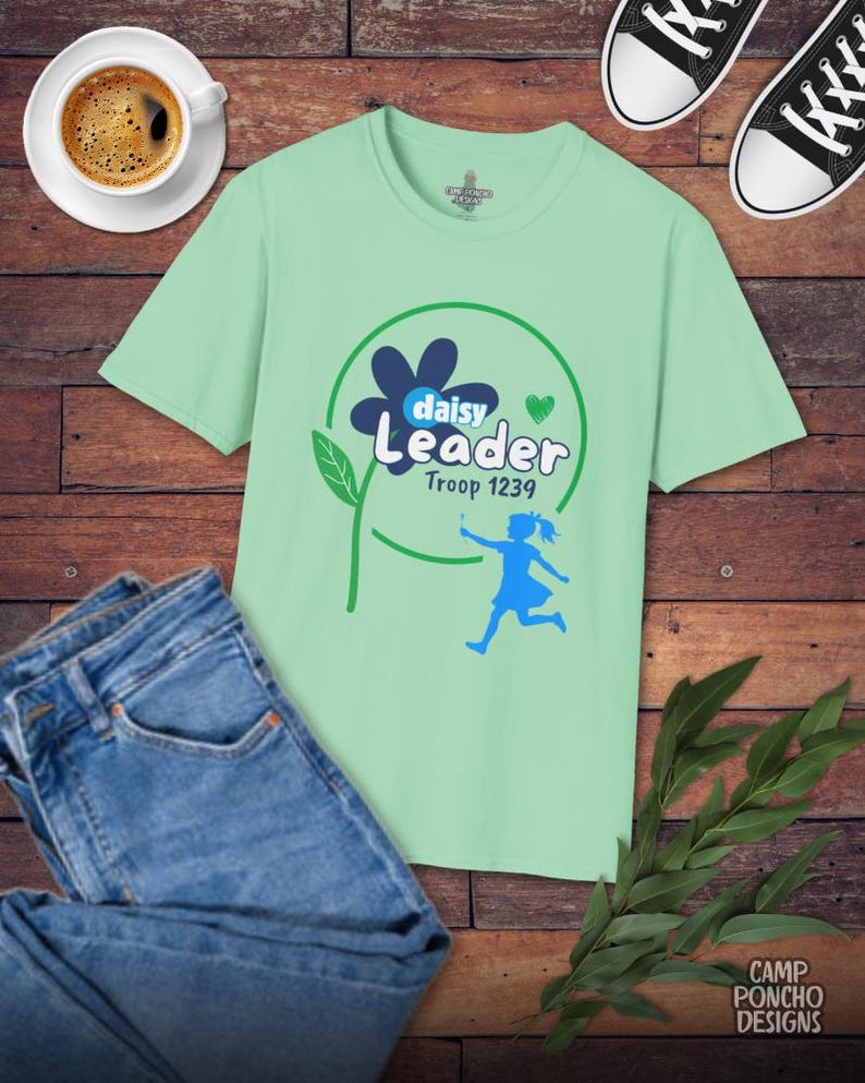 May include: A light green t-shirt with a graphic that says "Daisy Leader Troop 1239" with a flower and a running girl. The shirt is paired with blue jeans, black sneakers, and a cup of coffee on a wooden surface.