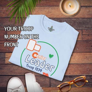 May include: A light blue t-shirt with the text "Senior Leader Troop 1239" in a green circle. The shirt is on a wooden surface with a lit candle, a pair of white sneakers, and tortoise shell glasses.