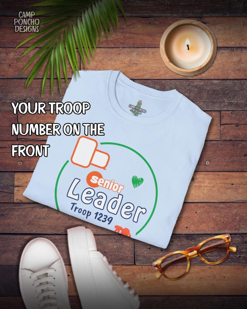 May include: A light blue t-shirt with the text "Senior Leader Troop 1239" in a green circle. The shirt is on a wooden surface with a lit candle, a pair of white sneakers, and tortoise shell glasses.