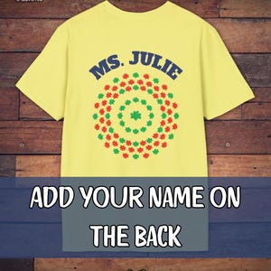May include: Yellow t-shirt with the text "MS. JULIE" in blue, above a circular design of red and green shapes. The bottom of the shirt has the text "ADD YOUR NAME ON THE BACK". The Camp Poncho Designs logo is in the top left corner.