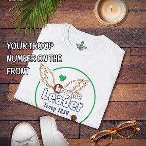 May include: White t-shirt with a green and brown graphic that says "Brownie Leader Troop 1239". The shirt also has the text "Your Troop Number on the Front". The shirt is on a wooden surface with a candle, glasses, and shoes.