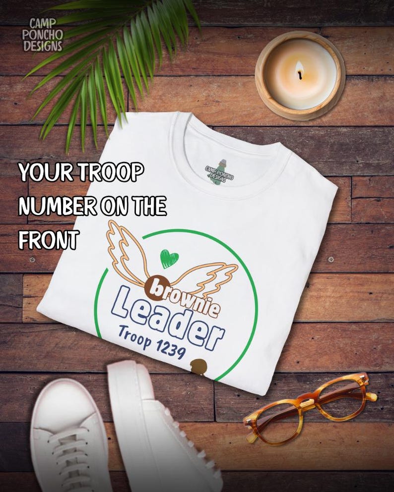 May include: White t-shirt with a green and brown graphic that says "Brownie Leader Troop 1239". The shirt also has the text "Your Troop Number on the Front". The shirt is on a wooden surface with a candle, glasses, and shoes.