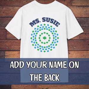May include: White t-shirt with the text "MS. SUSIE" in blue, above a circular design of green and blue shapes. The bottom of the shirt has the text "ADD YOUR NAME ON THE BACK". A green heart is at the bottom.