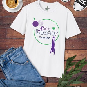 May include: White t-shirt with a green and purple graphic that says "Junior Leader Troop 1239" and a purple gymnast. The shirt is paired with blue jeans, black and white sneakers, and a cup of coffee on a wooden surface.