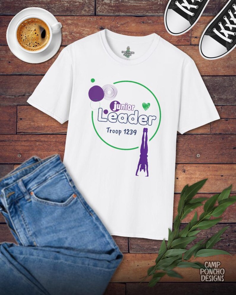 May include: White t-shirt with a green and purple graphic that says "Junior Leader Troop 1239" and a purple gymnast. The shirt is paired with blue jeans, black and white sneakers, and a cup of coffee on a wooden surface.
