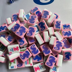 Puede incluir: Un conjunto de fichas de Mahjong rosas y blancas con dise&ntilde;os de caballitos de mar azules y caracteres chinos. Hay dados azules en la esquina superior izquierda. Las fichas son para un juego de mesa tradicional.