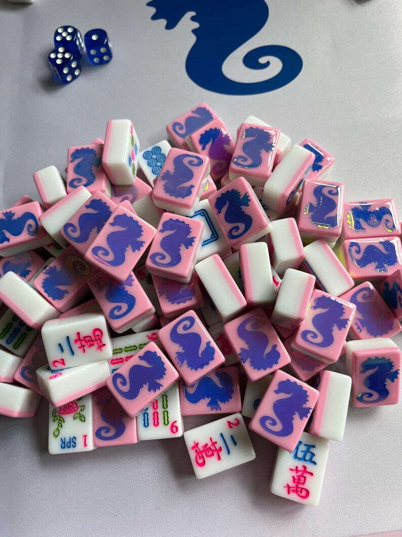 Puede incluir: Un conjunto de fichas de Mahjong rosas y blancas con dise&ntilde;os de caballitos de mar azules y caracteres chinos. Hay dados azules en la esquina superior izquierda. Las fichas son para un juego de mesa tradicional.