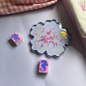 Puede incluir: Un posavasos decorativo con un dise&ntilde;o de caballito de mar y dos gomas de borrar rosas y blancas con gr&aacute;ficos de caballitos de mar. El posavasos es iridiscente con detalles en rosa y dorado. Una bolsa de mano crema con la palabra "MA" est&aacute; en el fondo.