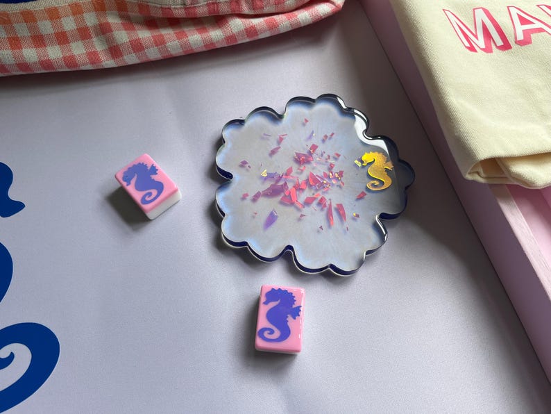 Puede incluir: Un posavasos decorativo con un dise&ntilde;o de caballito de mar y dos gomas de borrar rosas y blancas con gr&aacute;ficos de caballitos de mar. El posavasos es iridiscente con detalles en rosa y dorado. Una bolsa de mano crema con la palabra "MA" est&aacute; en el fondo.
