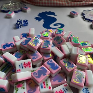 Puede incluir: Un conjunto de fichas de Mahjong rosas y blancas con dise&ntilde;os de caballitos de mar y caracteres chinos. Las fichas est&aacute;n dispuestas sobre una superficie blanca, con una silueta de caballito de mar azul en el fondo. Incluye dados azules y elementos decorativos.